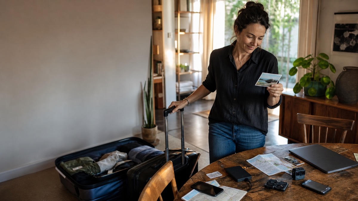 A woman at home looking at a travel photograph beside a phone, camera, cards and a suitcase.